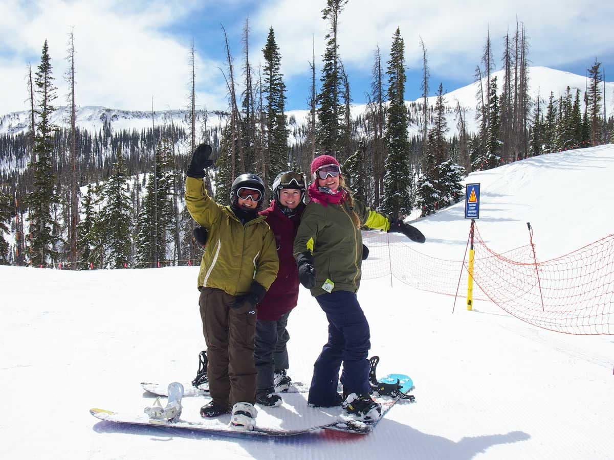 Three sowboarders pose for the camera at the top of a ski run with spindly evergreen trees and a semi-cloudy blue sky behind them
