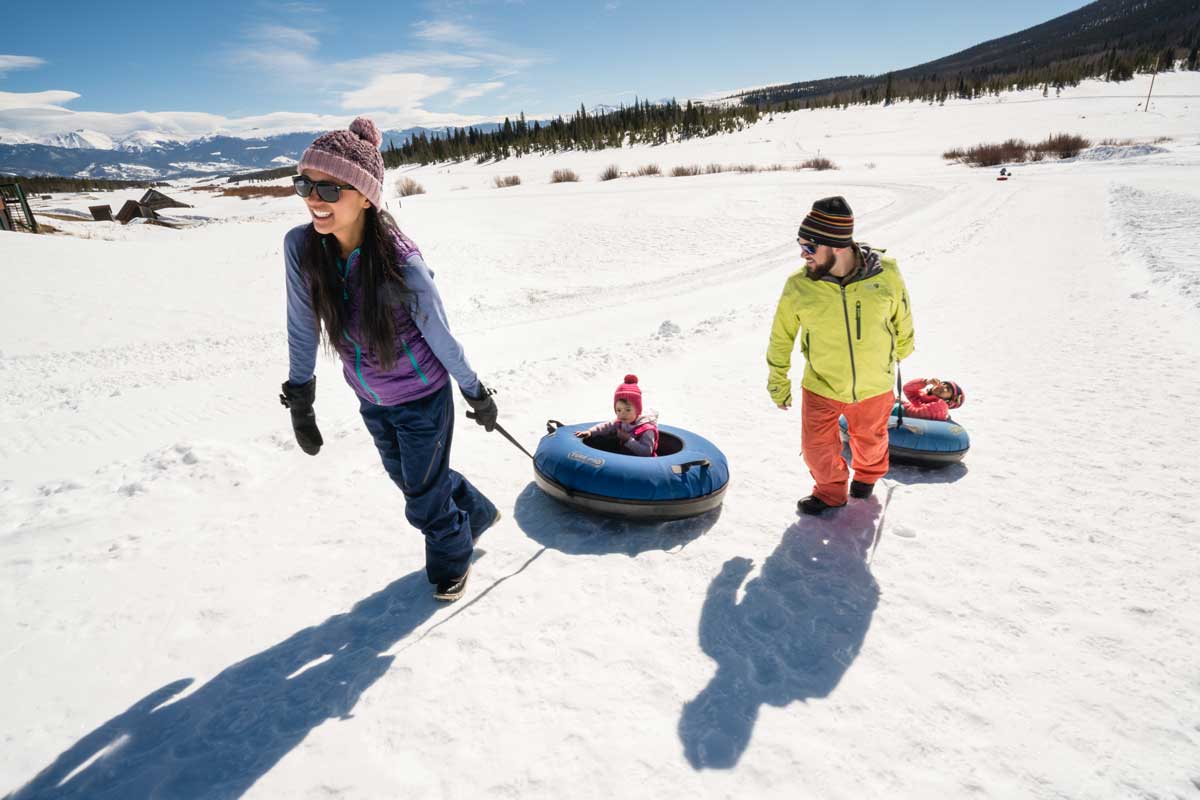 Two adults pull two children in snowtubs across a snowy landscape under a bright blue sky