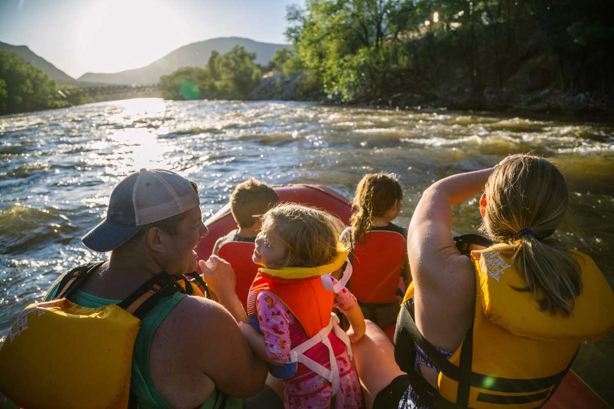 Two adults and three children ride along a relatively calm stretch of river. They all wear lifejackets and the sun is setting over the river