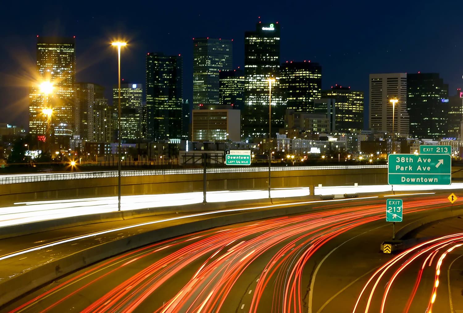 The skyscrapers of a Colorado city are lit and stand tall against a dark, night sky. In front of the city are curving highway road with green exits signs.