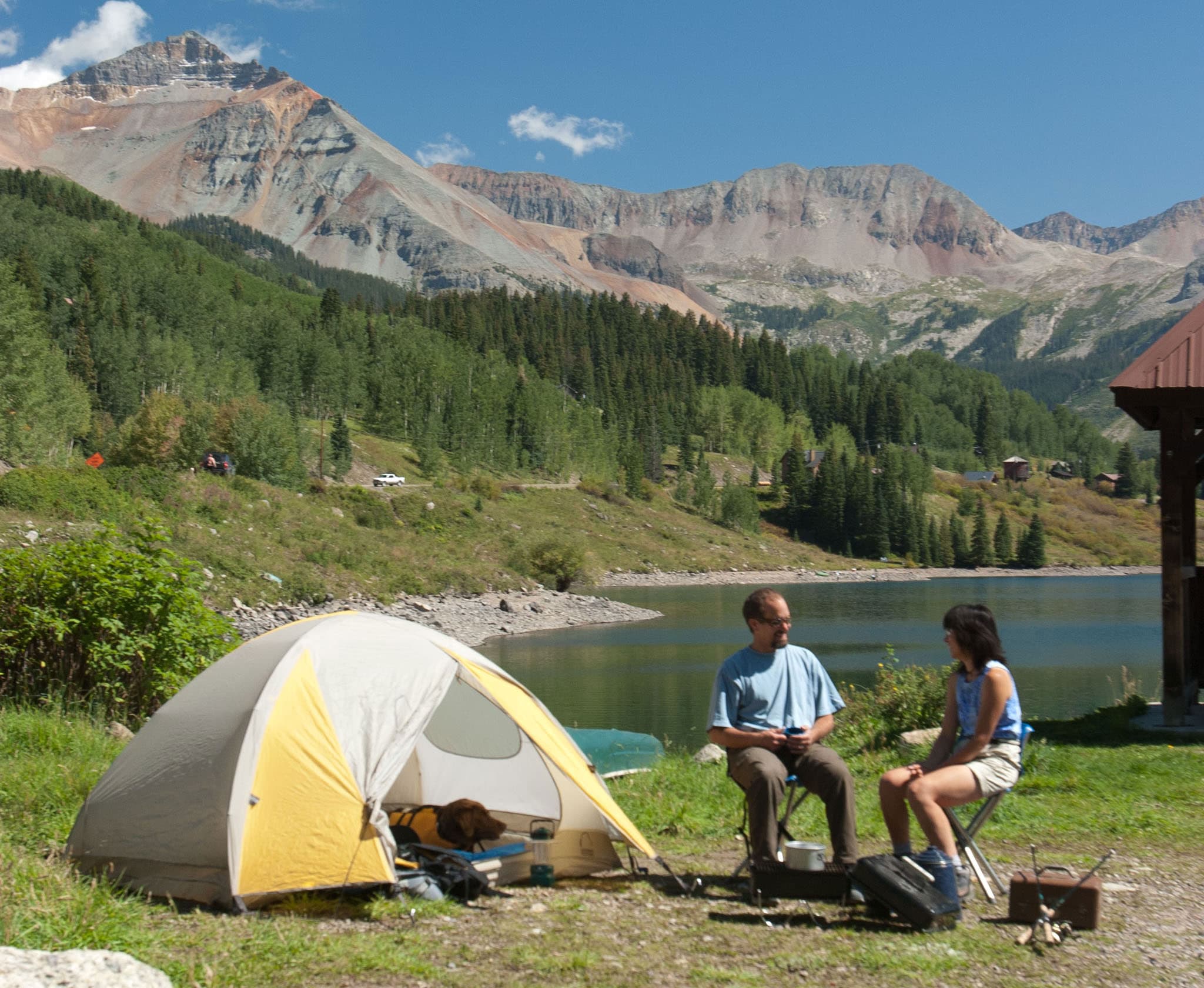 A couple sit on camp chairs next to a yellow tent that's next to a blue lake that's below a purpley-grey mountain ridge