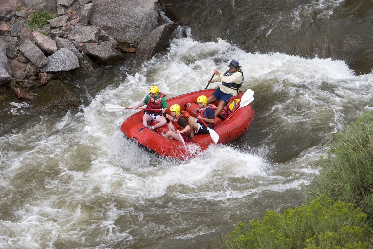 Inside a red raft, give paddlers in lifejackets dodge rocks and rapids while the guide in the back tries to steer them safely