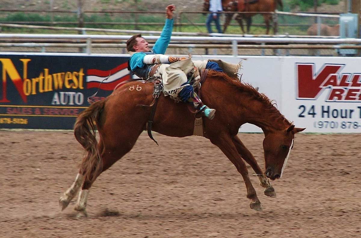 A cowpoke in a vibrant, pastel-blue long-sleeve shirt and chaps rides a bucking brown and white horse at the Meeker Range Call Rodeo.