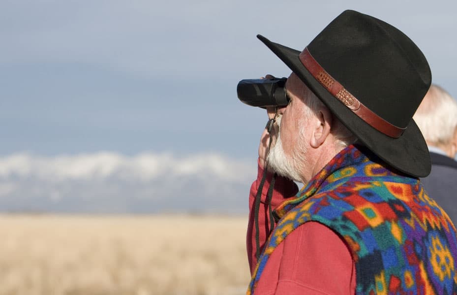 A person wearing a red shirt and fleece vest looks through a pair of black binoculars to watch for birds at Monte Vista's Crane Festival in Colorado.