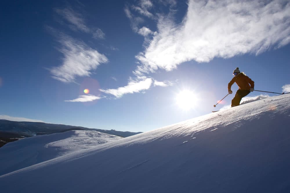 A skier shoots down a slope under a blue sky with a bright sun lighting up the scene.