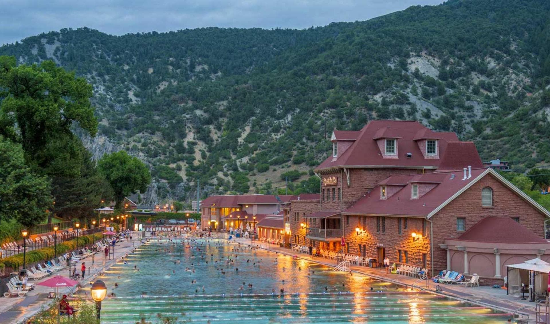 The massive pool of Glenwood Hot Springs with lots of people's heads bob in the water in the middle of the image. To the left green-leafed trees sit near a fence with pool loungers. On the right, the red-roofed, stone historic Glenwood Hot Springs building sits beneath a green tree covered mountain.