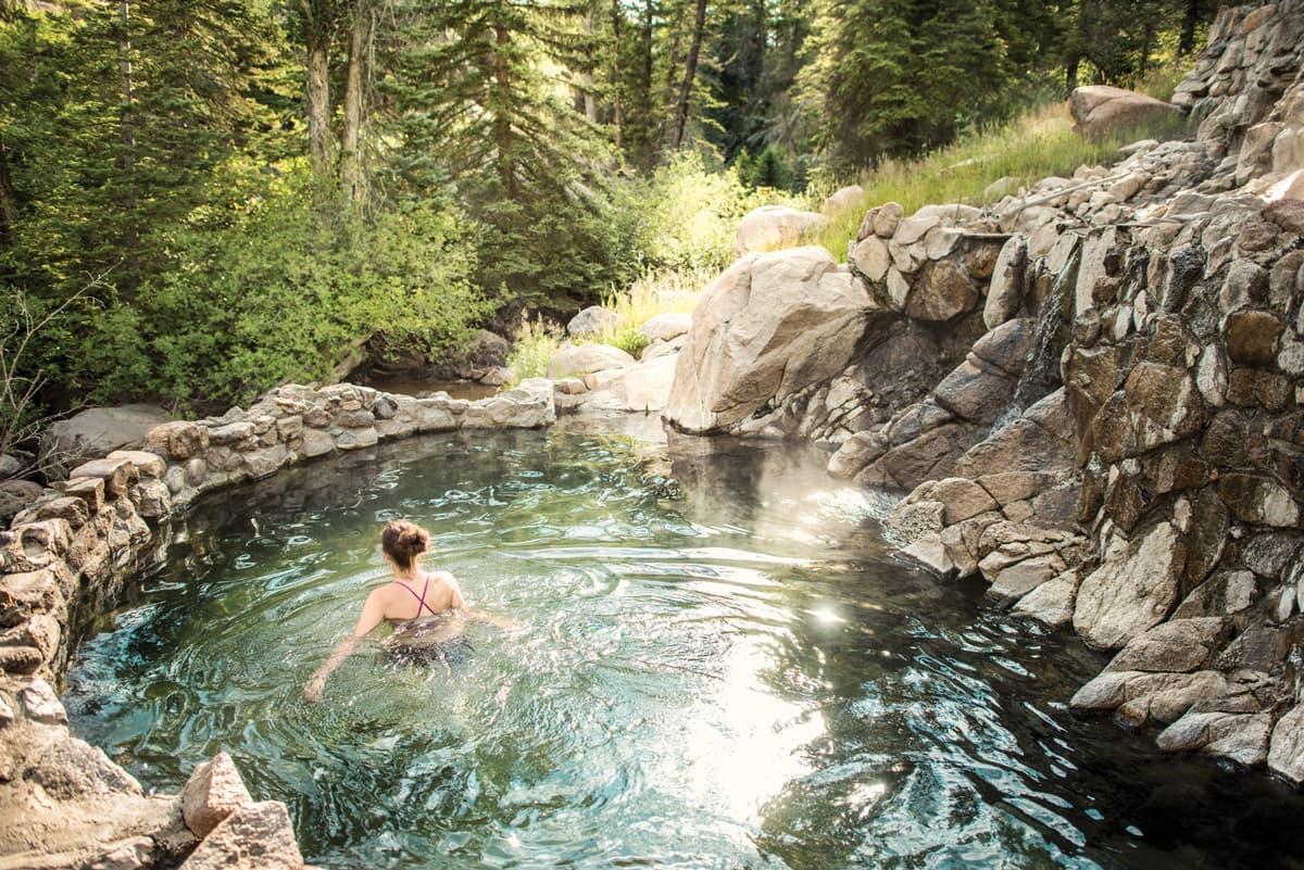 A person wades through a rippling pool at Strawberry Hot Springs Park. The pool is edged by a grey-stone wall and on the right a pile of stones leads into the pool. There are green-leafed trees in the background.