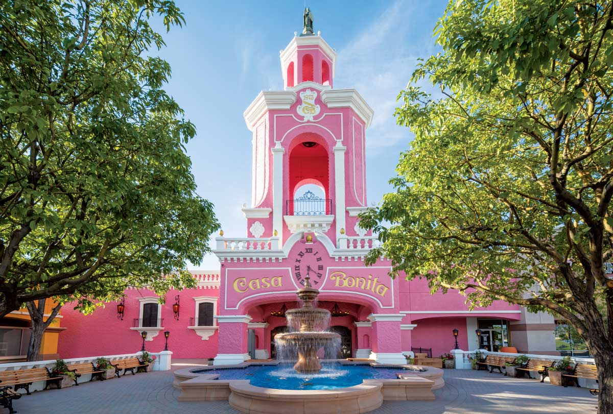 A bright-pink faux adobe building in Lakewood towers above a courtyard lined with wood benches and trees and centered around a flowing, two-tier fountain.