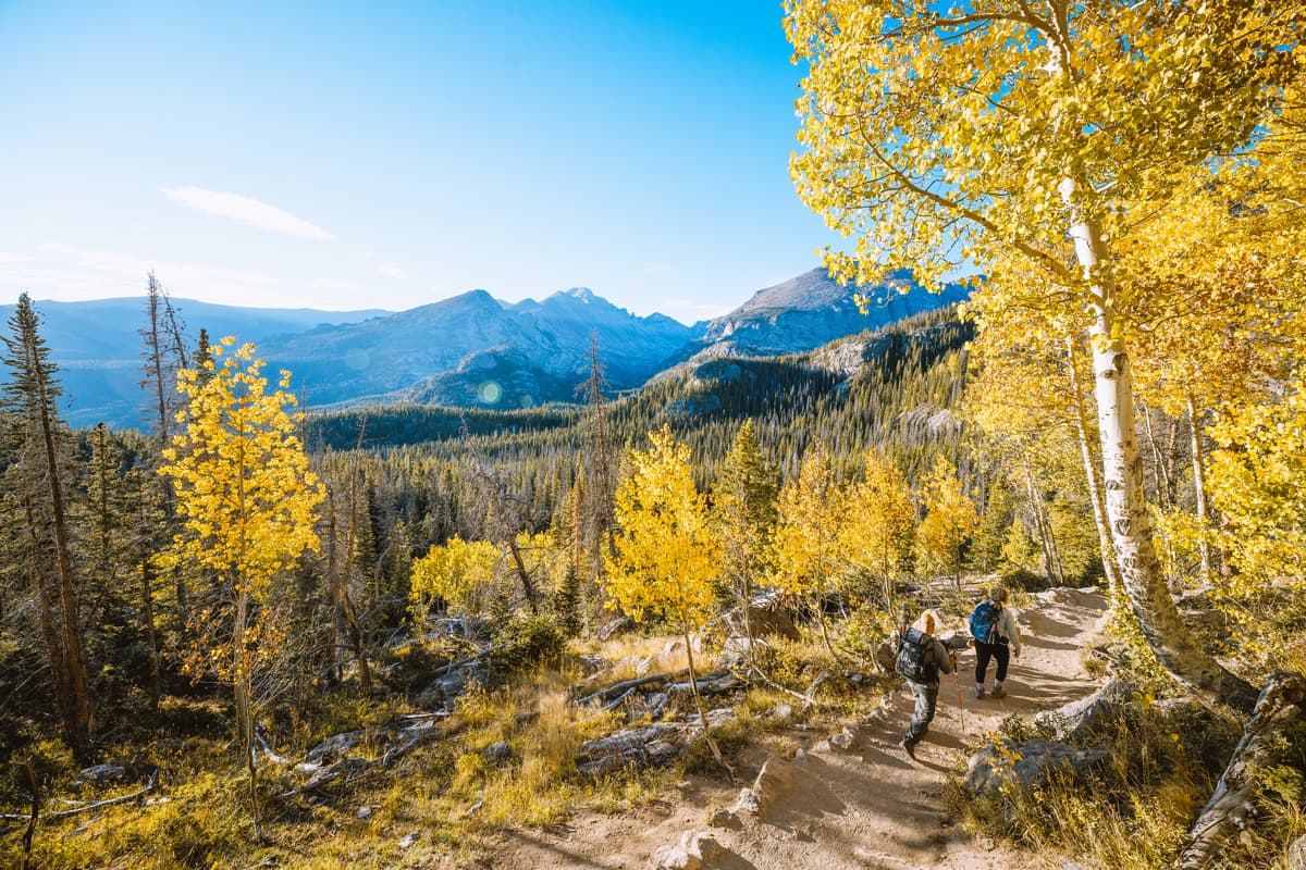 Two people hike on a trail lined with golden-leafed aspen trees with mountain views in Estes Park, Colorado.