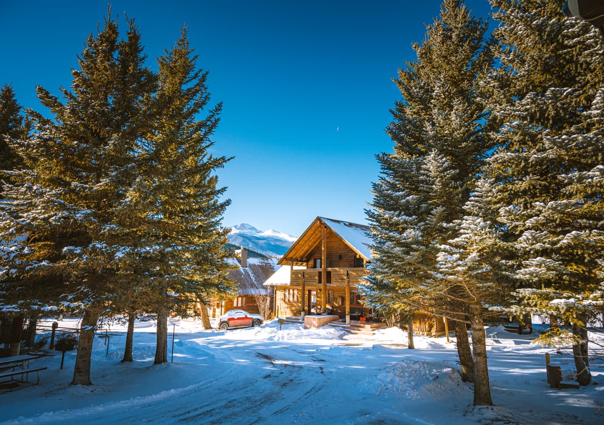 A snow-flocked cabin flanked by evergreens and a mountain backdrop delights at Rams Horn Village Resort in Estes Park, Colorado.
