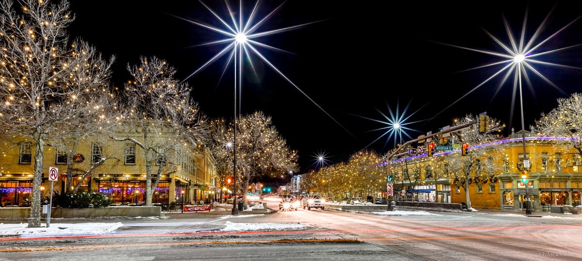 The snow-covered convergence of two streets in downtown Fort Collins on a winter's night. There are white twinkle lights in all of the trees and a couple cars are on the road.