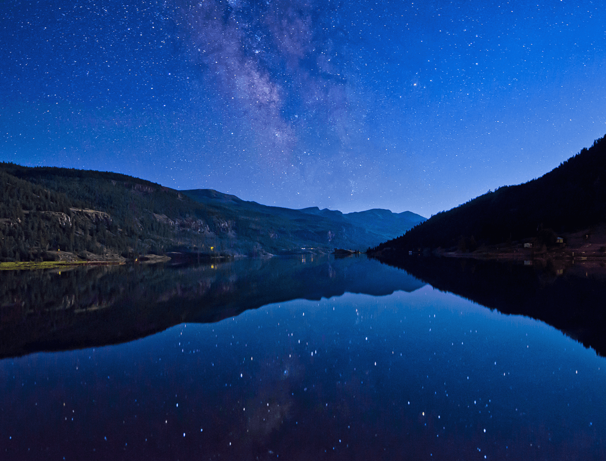 The night's sky with stars are reflected on the glassy surface of a mountain lake. On both sides evergreen trees run up to the shore with mountain peaks in the distance.