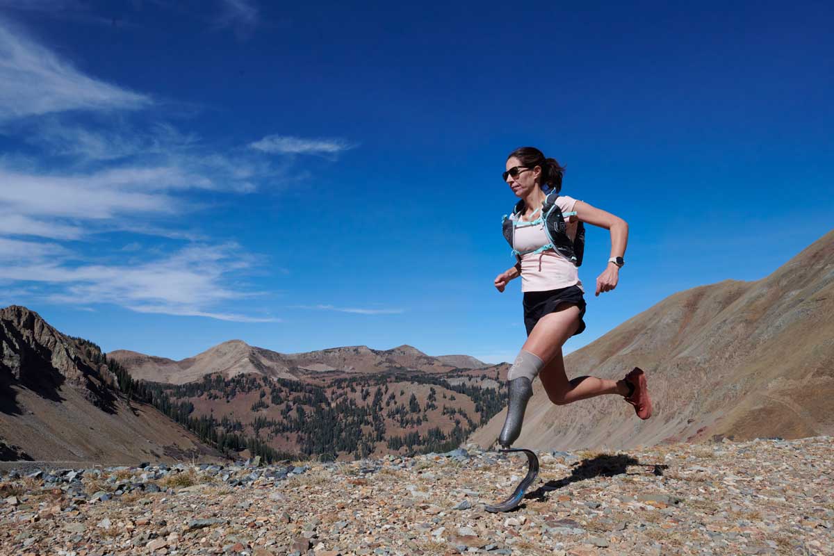 A person with a running blade and wearing a hydration vest runs across high-mountain terrain with Colorado peaks in the distance.