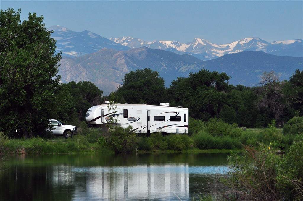 A white RV is parked next to a lake. Behind it is a lush grove of trees and snowtopped mountan peaks