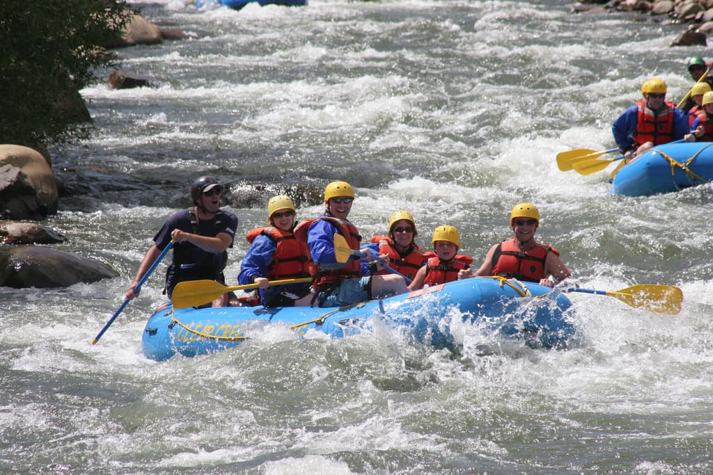 Five people in yellow helmets and red jackets paddle through fierce rapids while a guide at the back of the blue boat shouts instructions at them. They all appear to be smiling