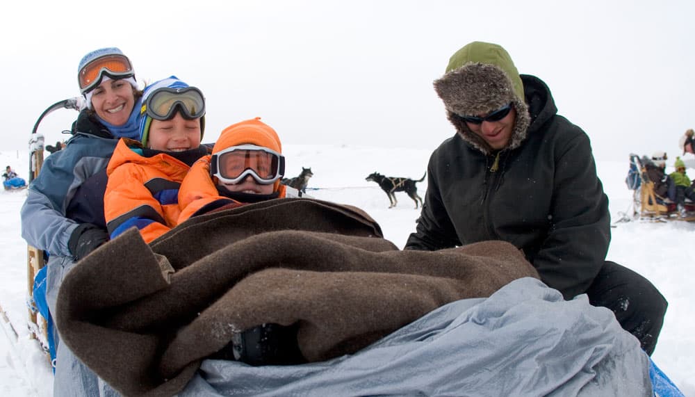 An adult and two kids wear goggles and get bundled up into a sled with layers of warm blankets in preparation for a dog-sledding adventure near Edwards, Colorado.