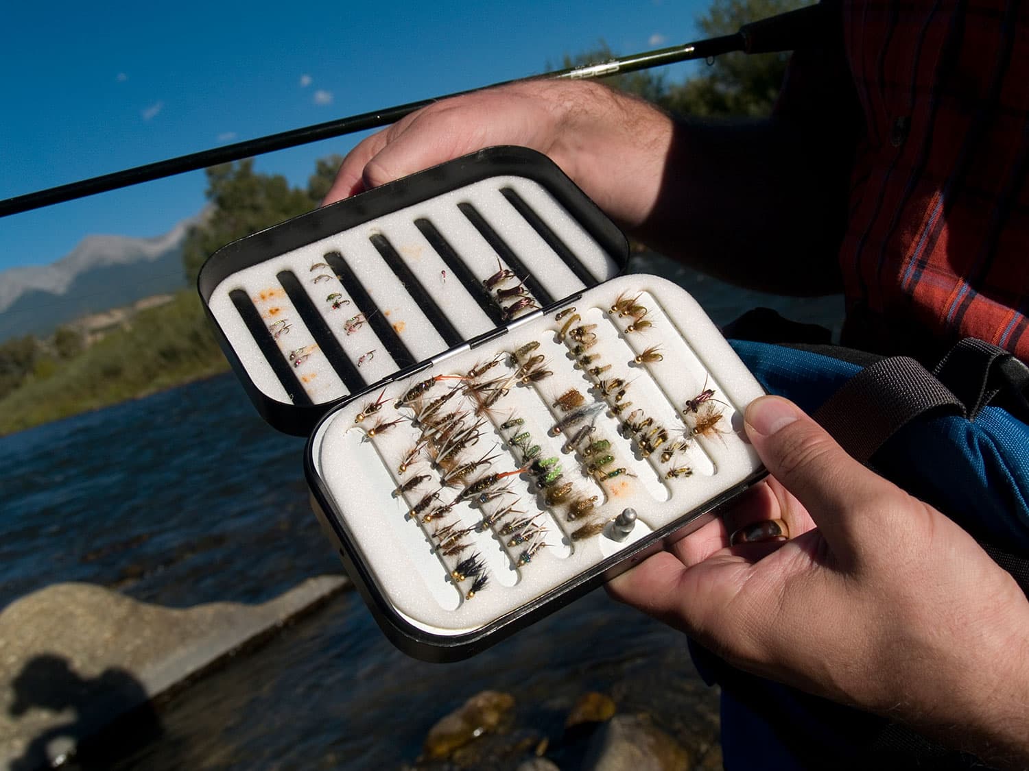 A person opens a tin lined with small, insect-like lures set on a padded lining. They are preparing to cast their link on the Arkansas River near Salida, Colorado.