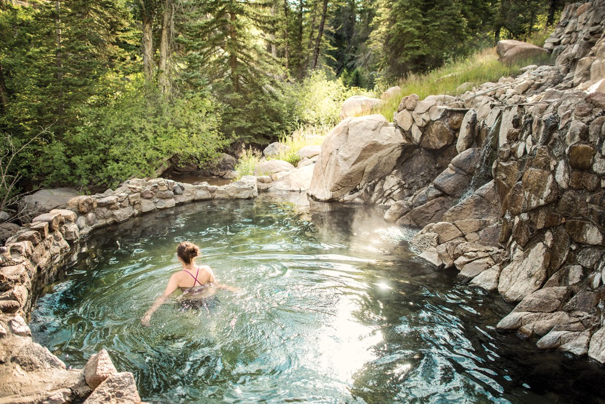 A person wades through a rippling pool at Strawberry Hot Springs Park. The pool is edged by a grey-stone wall and on the right a pile of stones leads into the pool. There are green-leafed trees in the background.