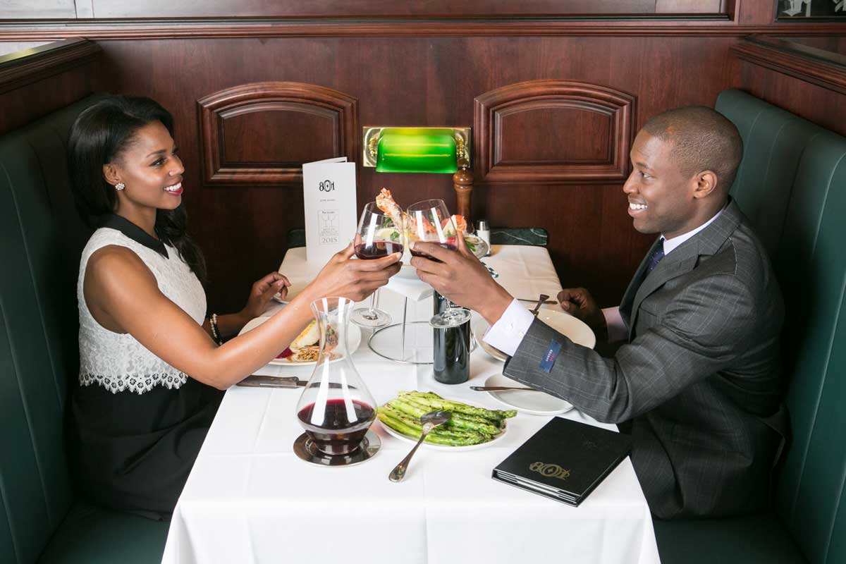 Two people sit across from each other in an elegant with rich wooden walls and a white table cloth at Cherry Creek North. They clink glasses of red wine.