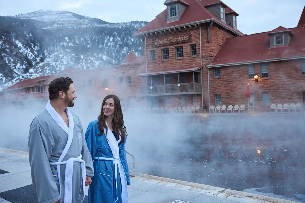 Two people in robes walk beside the steamy waters of Glenwood Hot Springs in winter