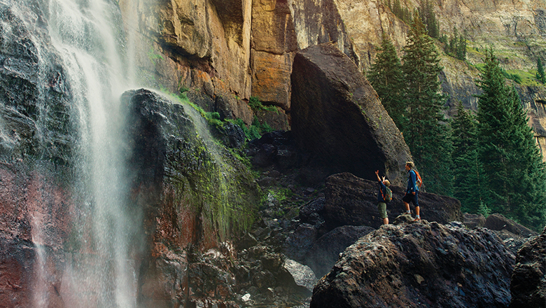 Two children raise their hands excitedly in front of rushing water of Bridal Veil Falls