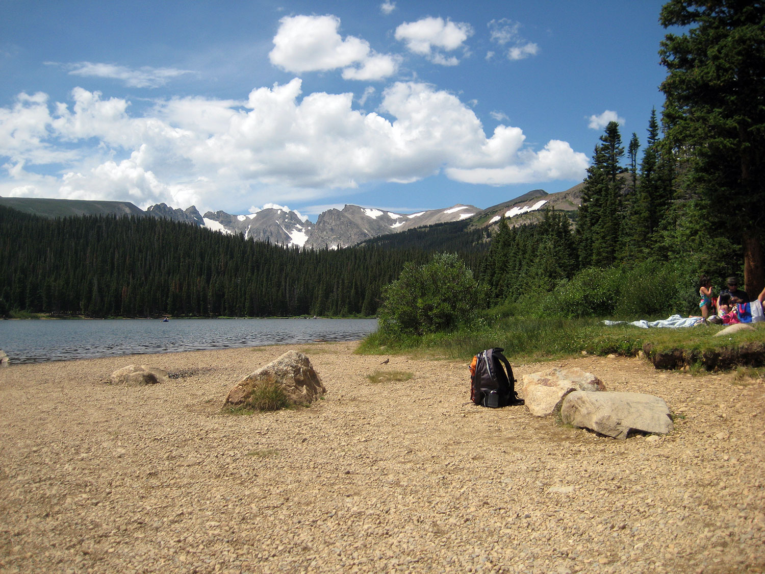 A group of picnickers sit on a blanket on the sandy shore of a blue mountain lake