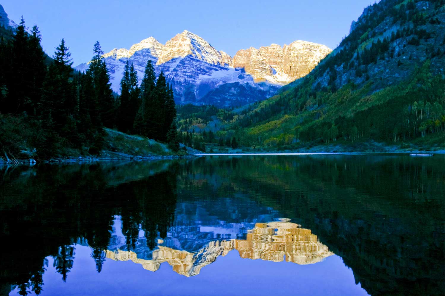 Maroon Bells peaks reflect on the surface of a lake below with trees and hillsides in the foreground.