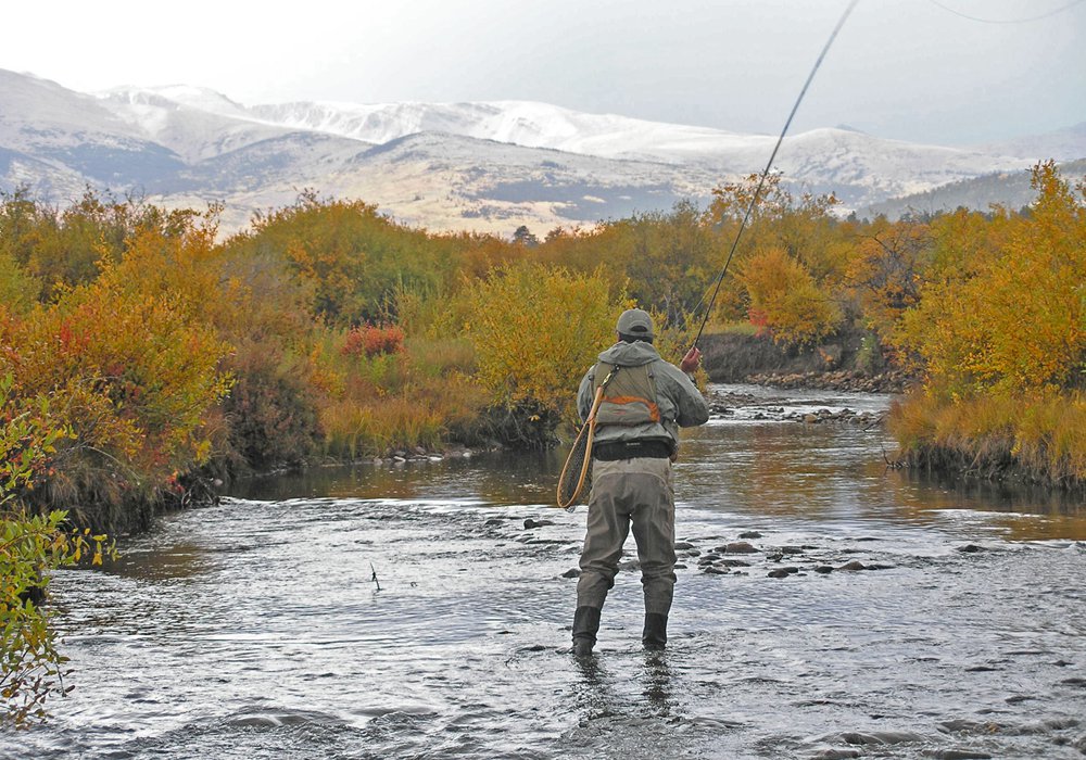 A flyfisher in waders casts his line from the middle of a river, surrounded on both sides by the changing orange and yellow leaves of fall
