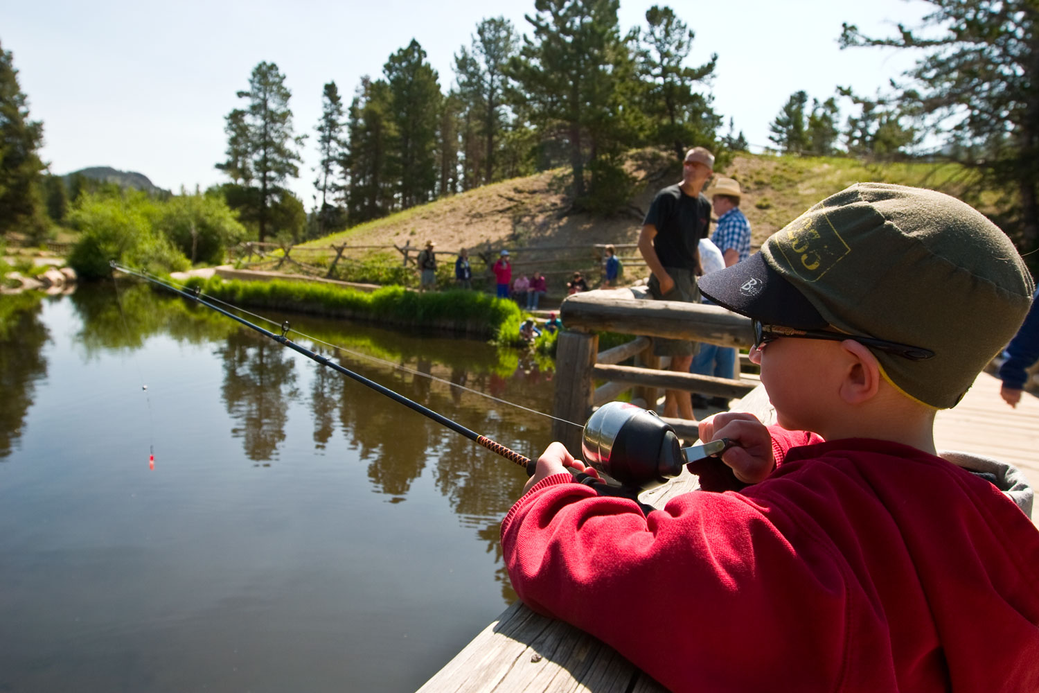 A boy in a green hat and red sweatshirts holds his fishing pole over a bridge, dropping his line into a still creek below