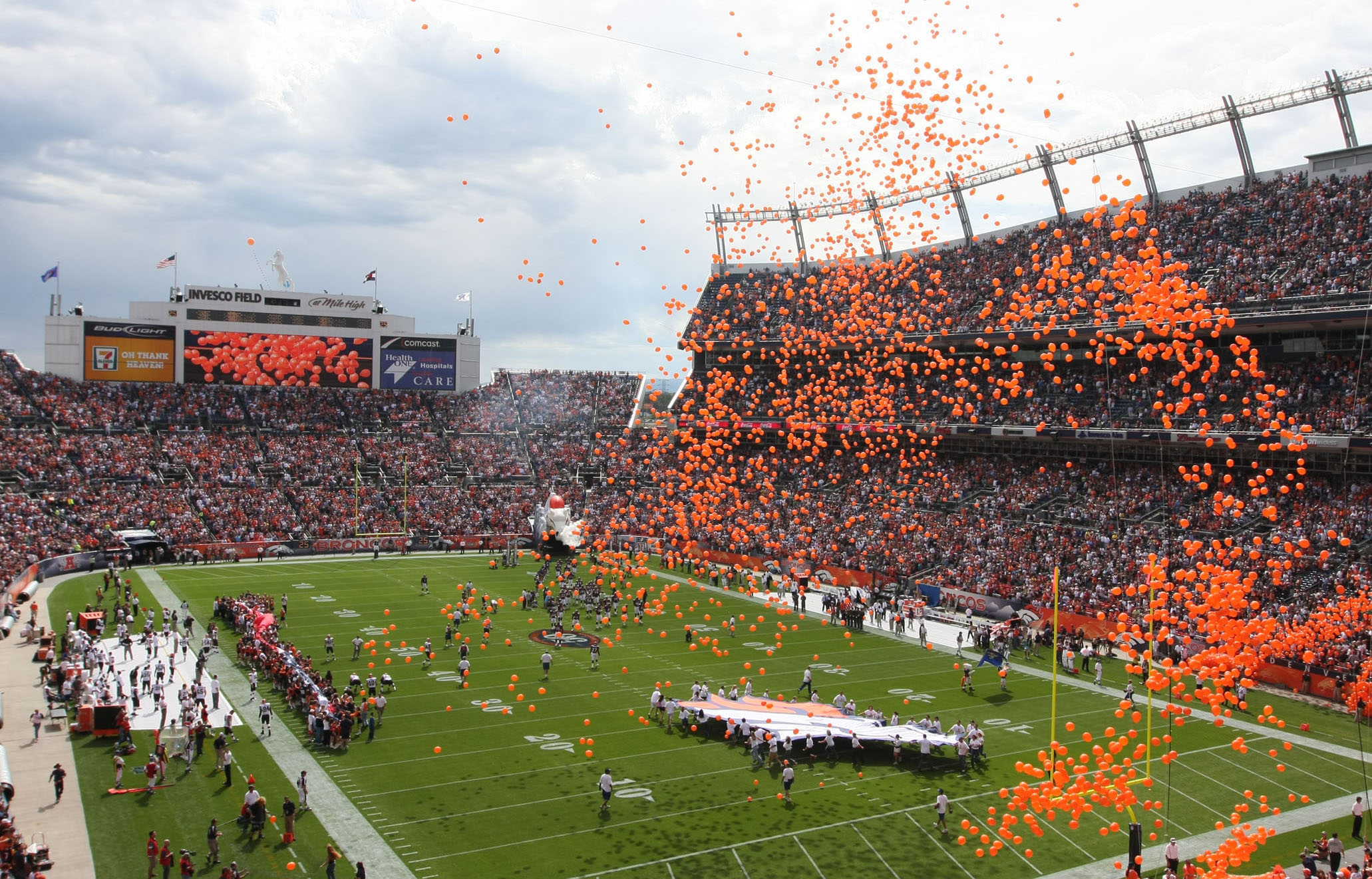 Orange balloons float through the air at the Denver Bronco's football stadium. People on the field hold out a giant Broncos logo.