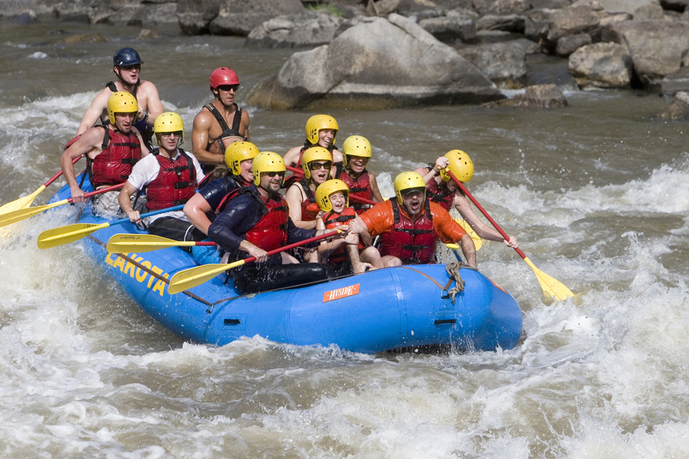 12 people in yellow helmets and red life jackets hold yellow paddles and ride the river waves in an inflatable blue boat
