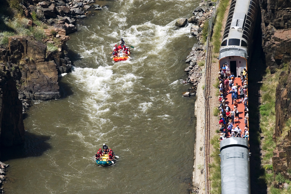 An aerial view of two rafts filled with paddlers passing by a train on the banks of the river