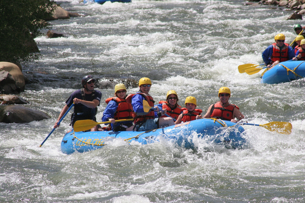 Five people in yellow helmets and red jackets paddle through fierce rapids while a guide at the back of the blue boat shouts instructions at them. They all appear to be smiling