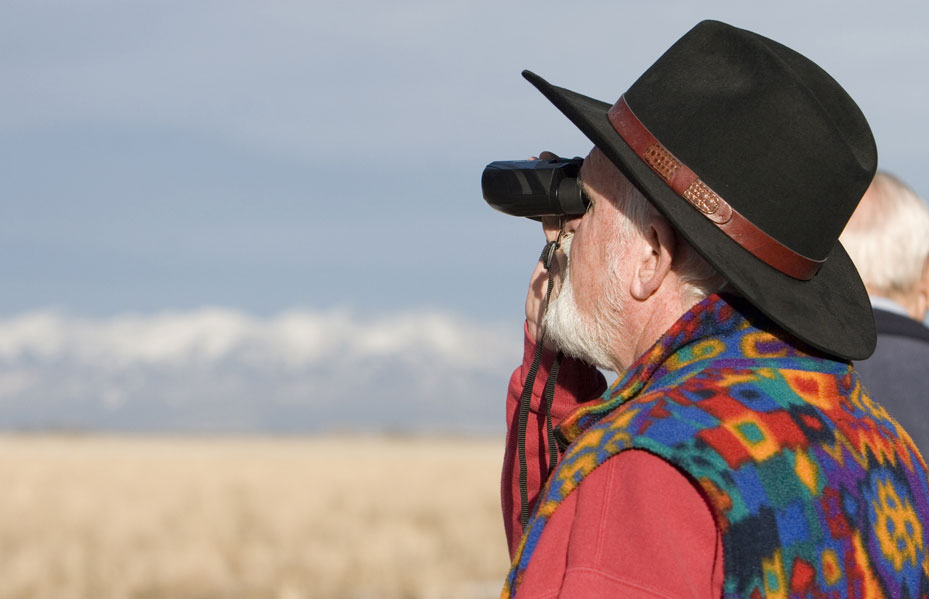 A person wearing a red shirt and fleece vest looks through a pair of black binoculars to watch for birds at Monte Vista's Crane Festival in Colorado.