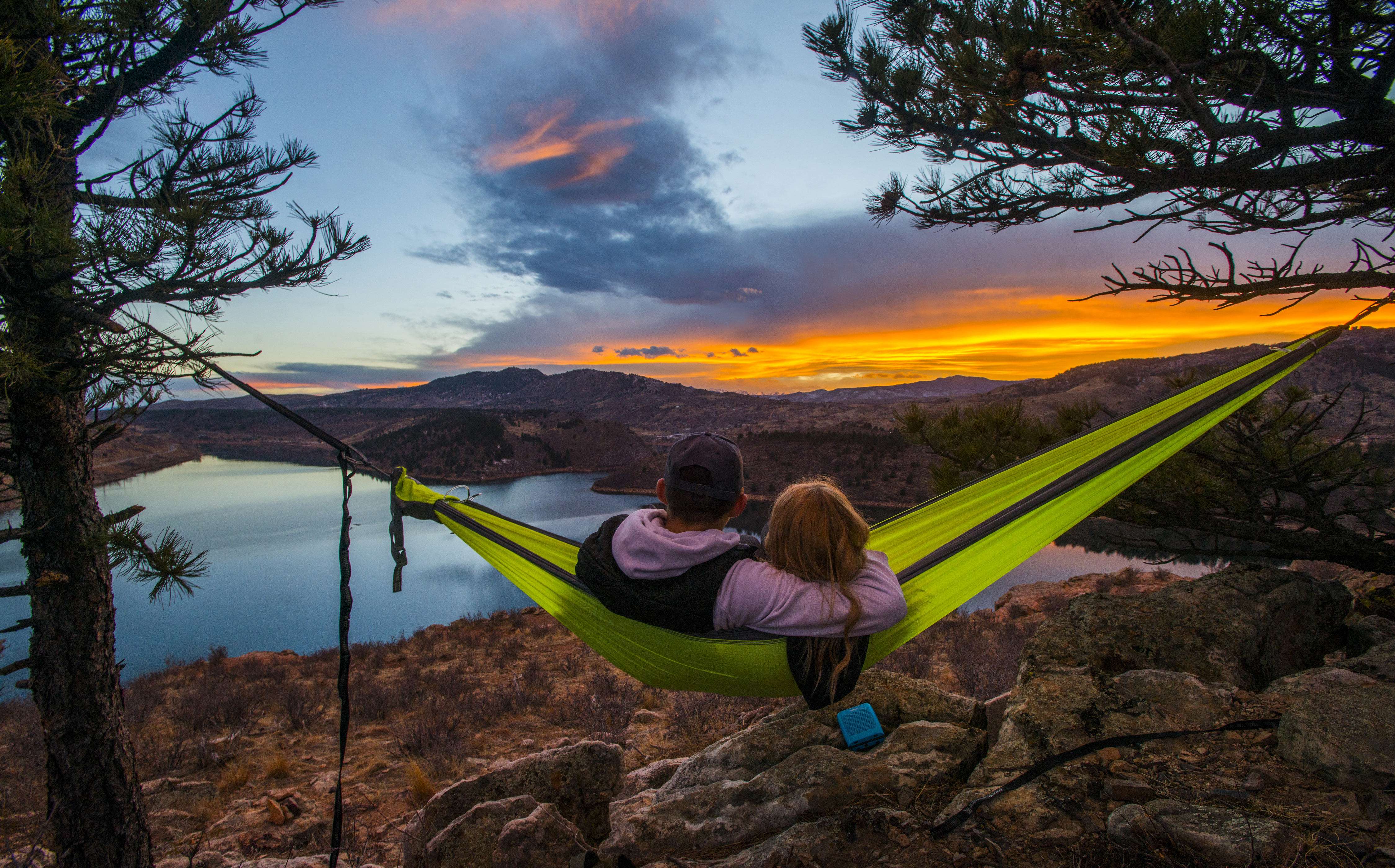 A couple in a lime-green hammock, that's strung up between two pine trees above a rocky outcropping look out onto Horsetooth Reservoir at sunset. In the distance a mountain meets an orange sky that's tinged with blue.