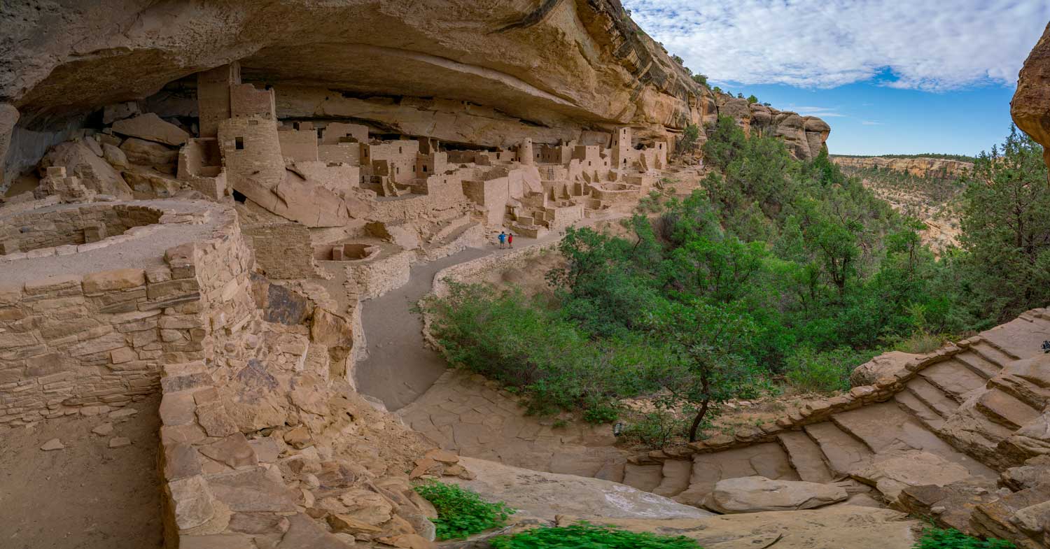Cliff formations of Mesa Verde National Park stretch to more than halfway through the middle of the photo, surrounded by bright-green trees. All of this is underneath a bright-blue sky sprinkled with white clouds.