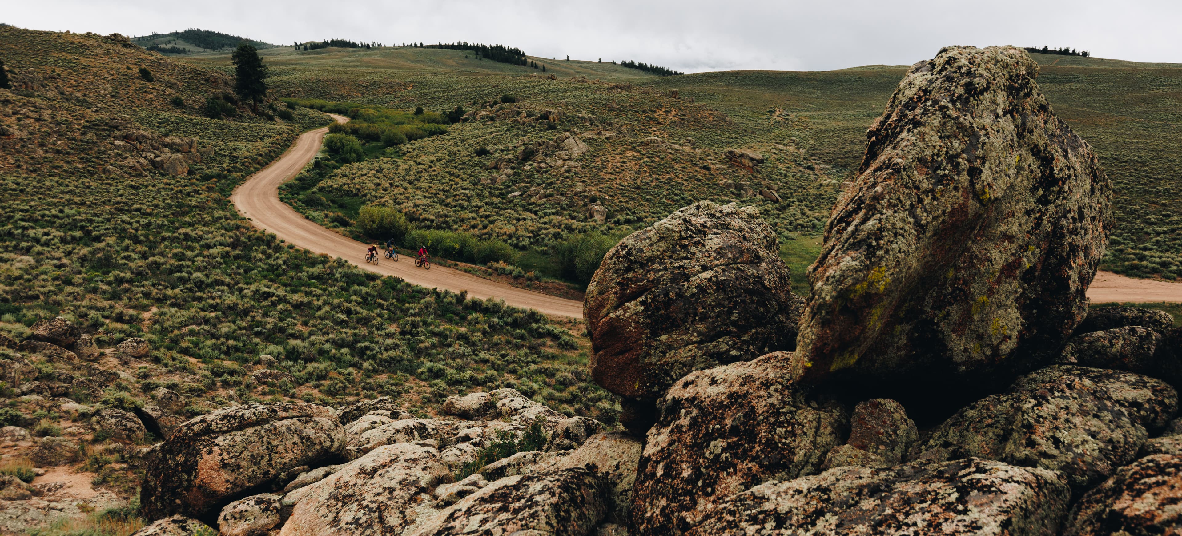 People on mountain bikes ride a trail through green hills and rocky terrain in Gunnison Crested Butte in Colorado