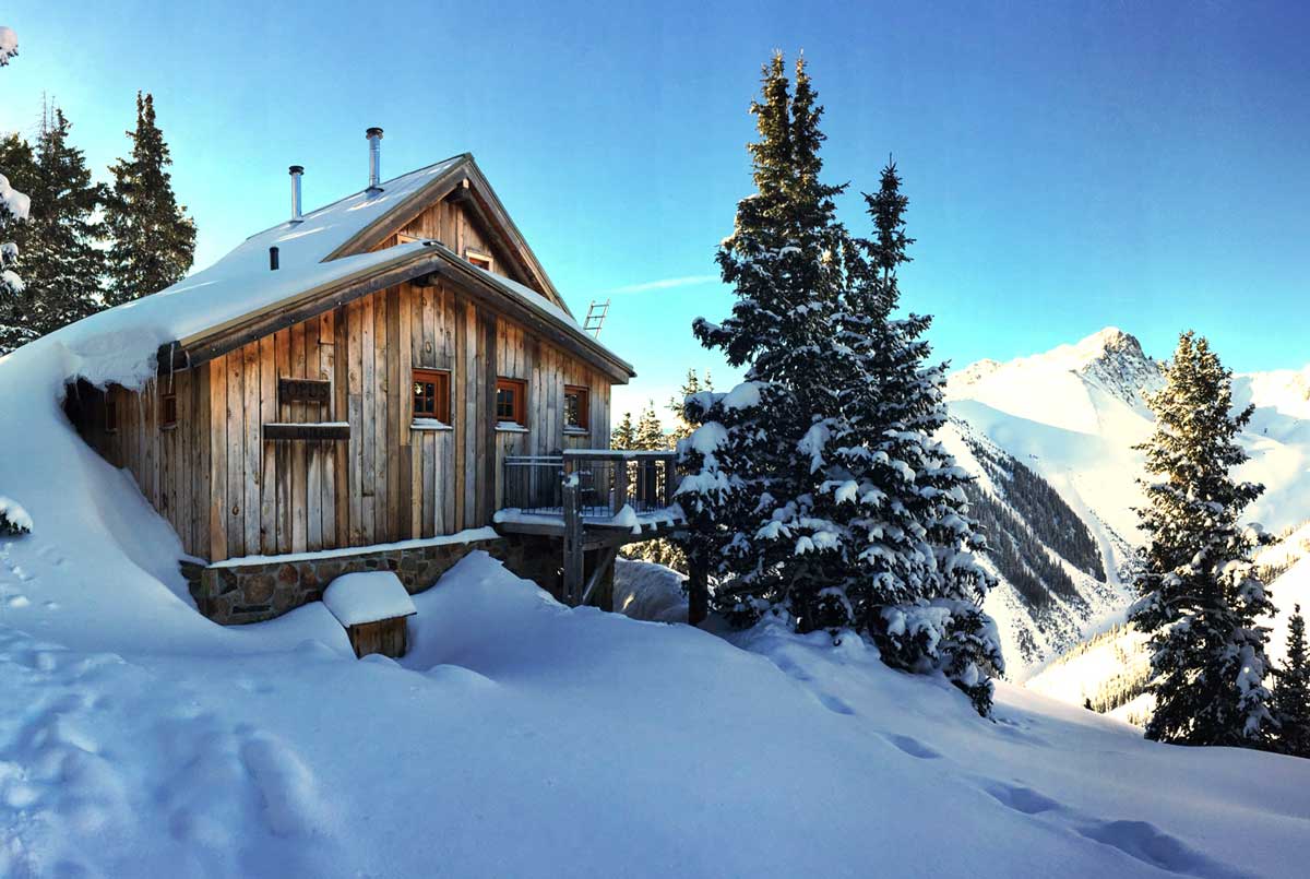 The wood-paneled Opus Hut near Silverton is covered in several feet of snow under a blue sky with mountain peaks in the Colorado background.