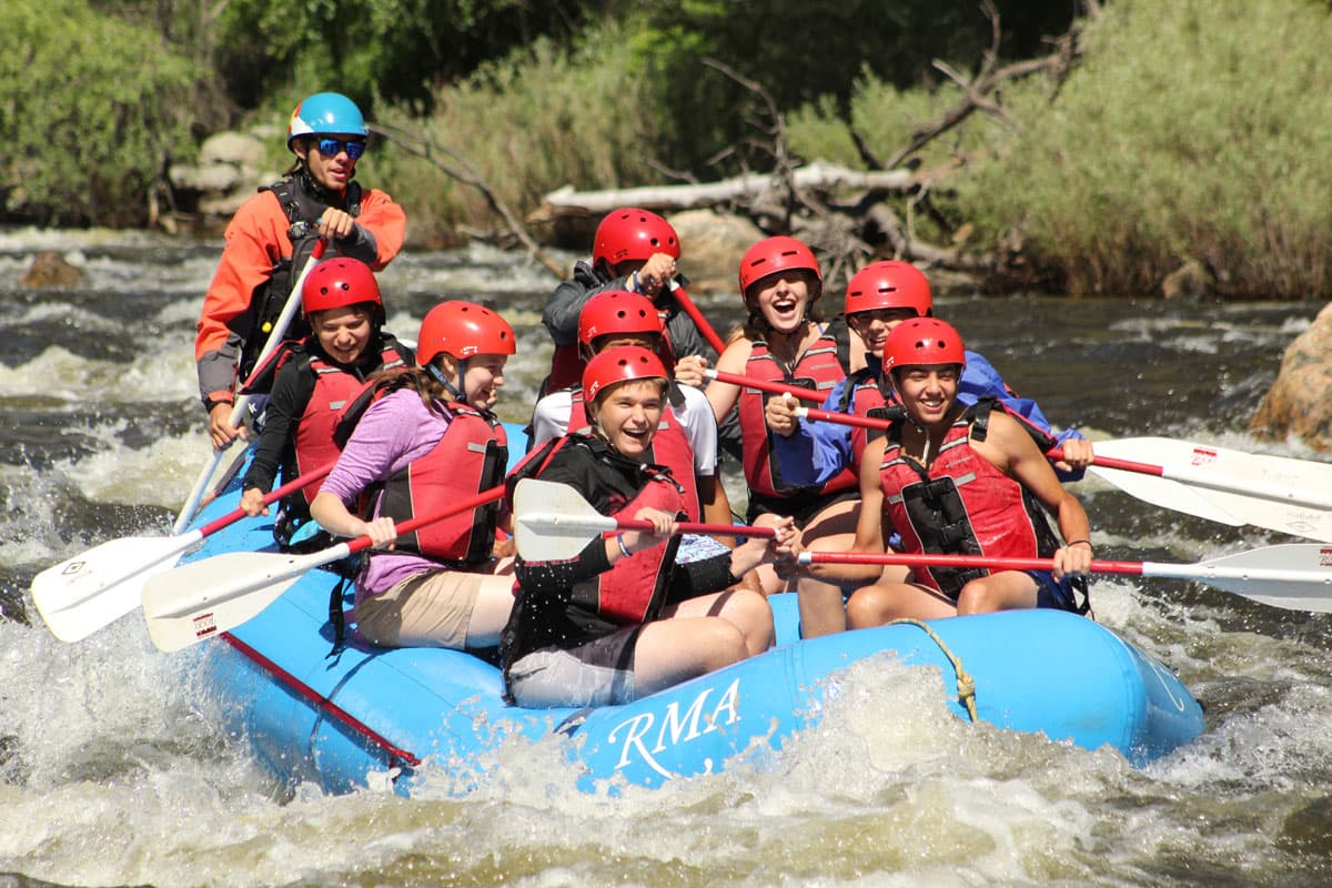 A group of red vest- and helmet-clad younger people sit in an inflated blue raft on the rolling whitewater waves of the Cache la Poudre River in Colorado. They are smiling and holding paddles in their hands while the instructor in the back of the raft guides them along the water. In the background are green plants and trees lining the river.