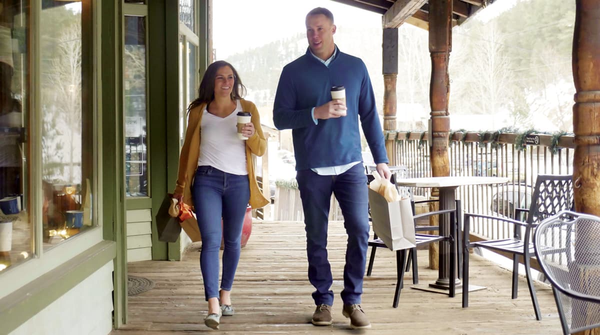 Two people are smiling, talking and holding coffees as they walk towards the camera, casually enjoying a sunny day in Downtown Evergreen on the iconic wooden boardwalk.