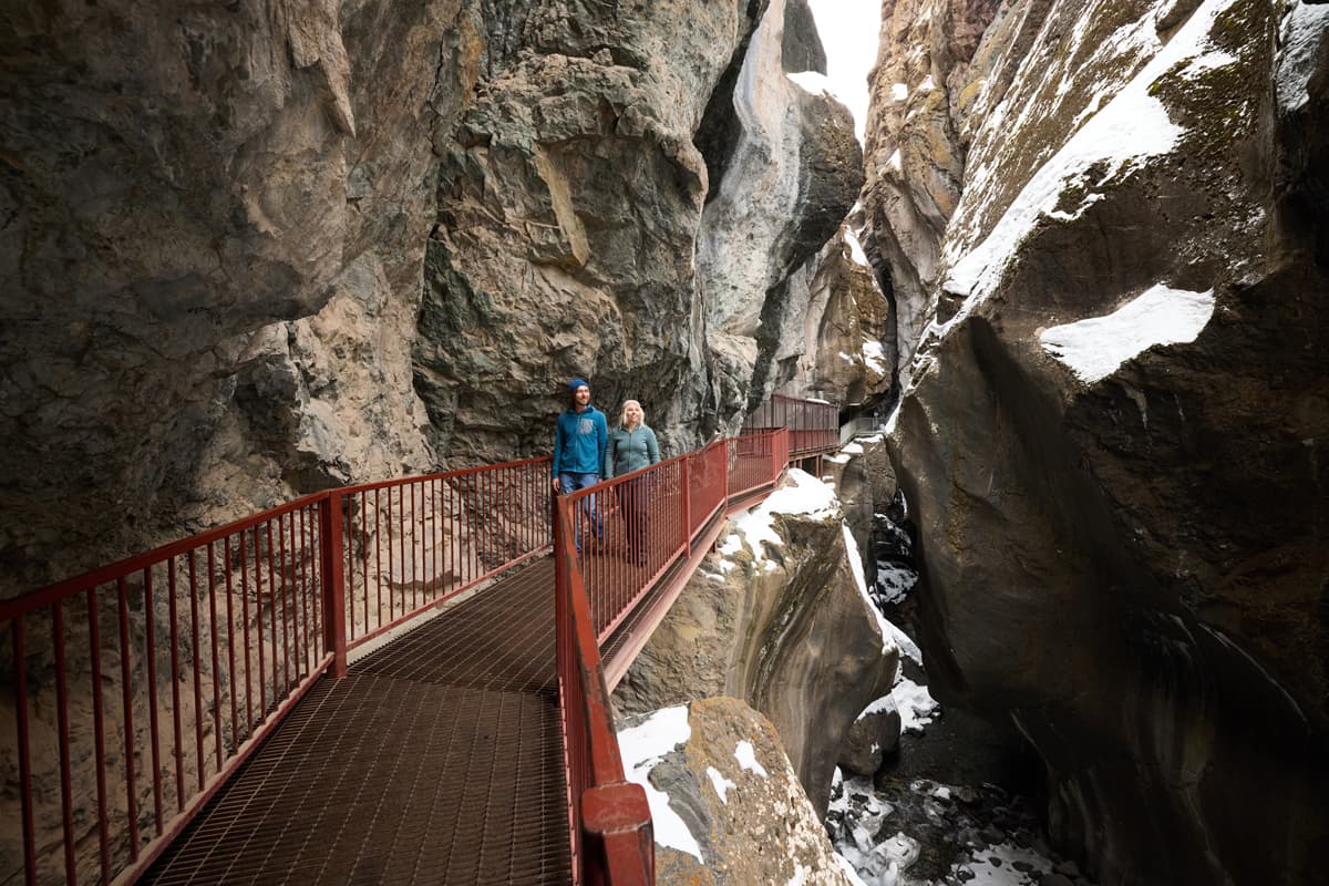 Two people walk along a bridge past snow covered rock walls at Box Cañon Falls in winter in Ouray, Colorado