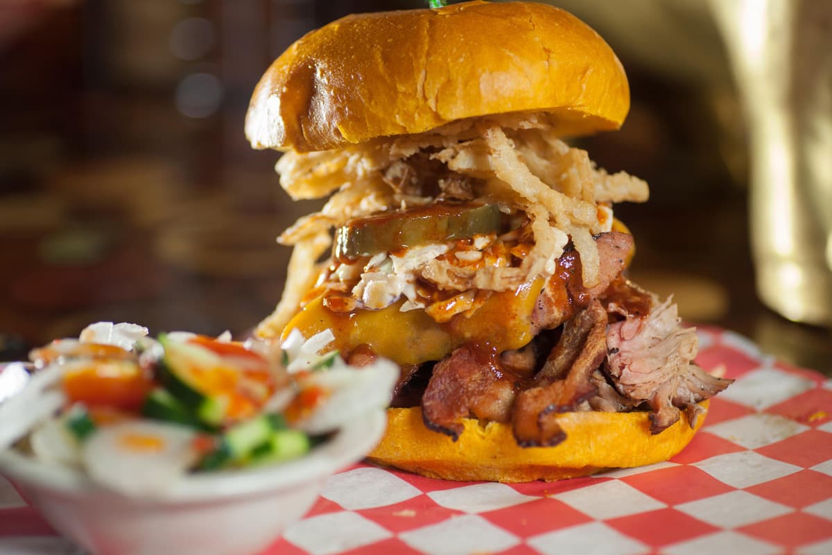 On a red and white checked placemat in Colorado Springs sits a roast beef sandwich stacked with pickles and onion rings on a brioche bun. In front of the sandwich is a small, white bowl filled with salad.