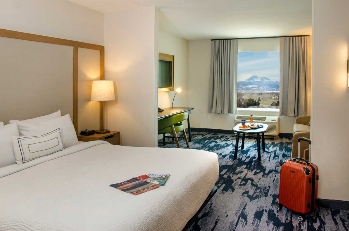 Guest room with one bed with white bedding, a desk with a green leather chair, a coffee table with food on it and an orange roller board suitcase at Fairfield Inn & Suites by Marriott Boulder Longmont