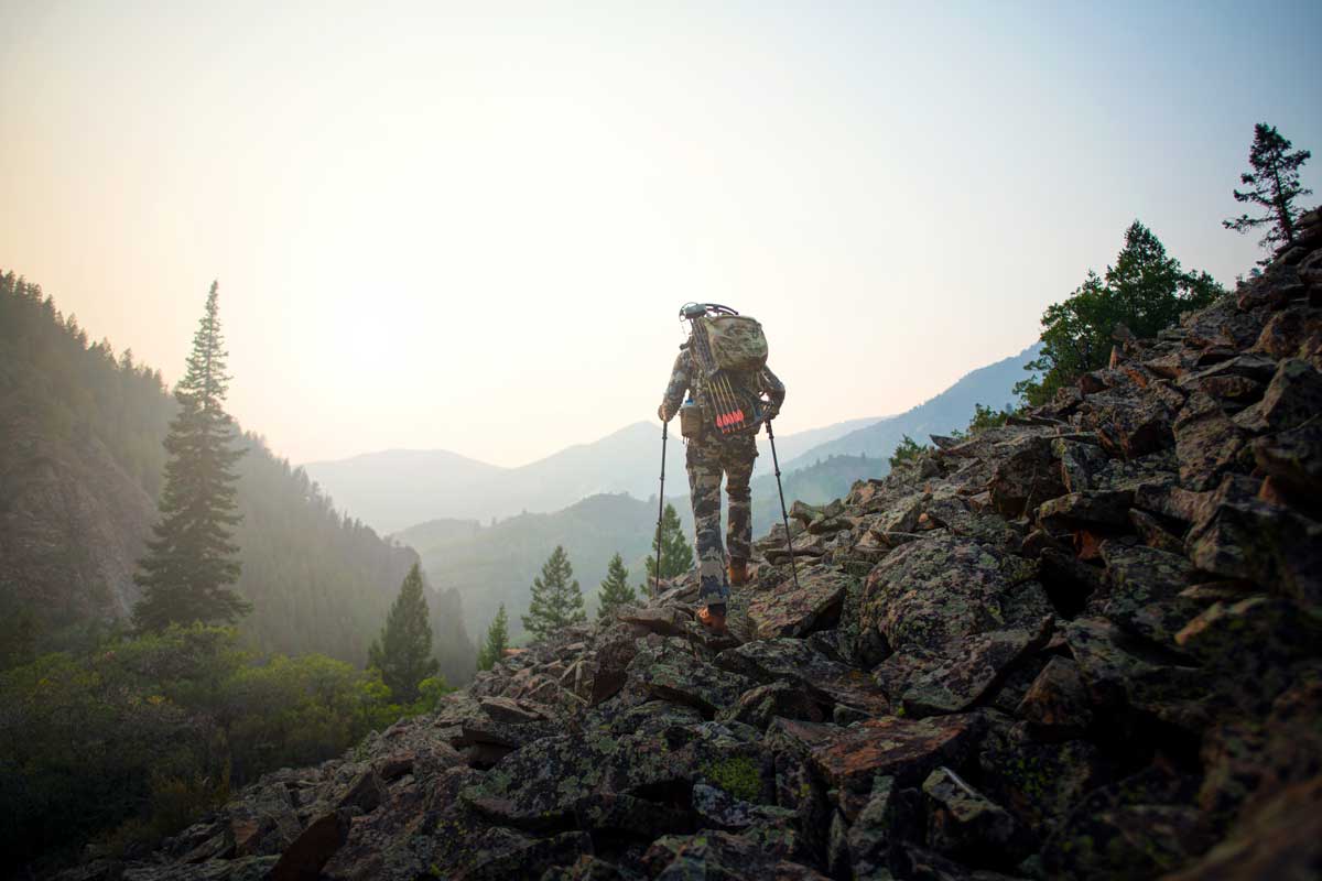 A bow hunter in a camo outfit uses walking sticks to navigate along a craggy area of rocks in the Colorado Rocky Mountains.