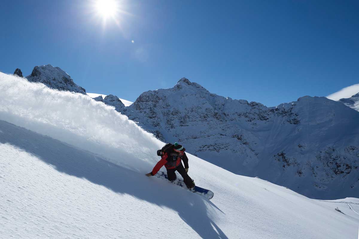 A snowboarder shreds fresh, untouched powder on a steep slope in Colorado. A wave of powder forms behind them.