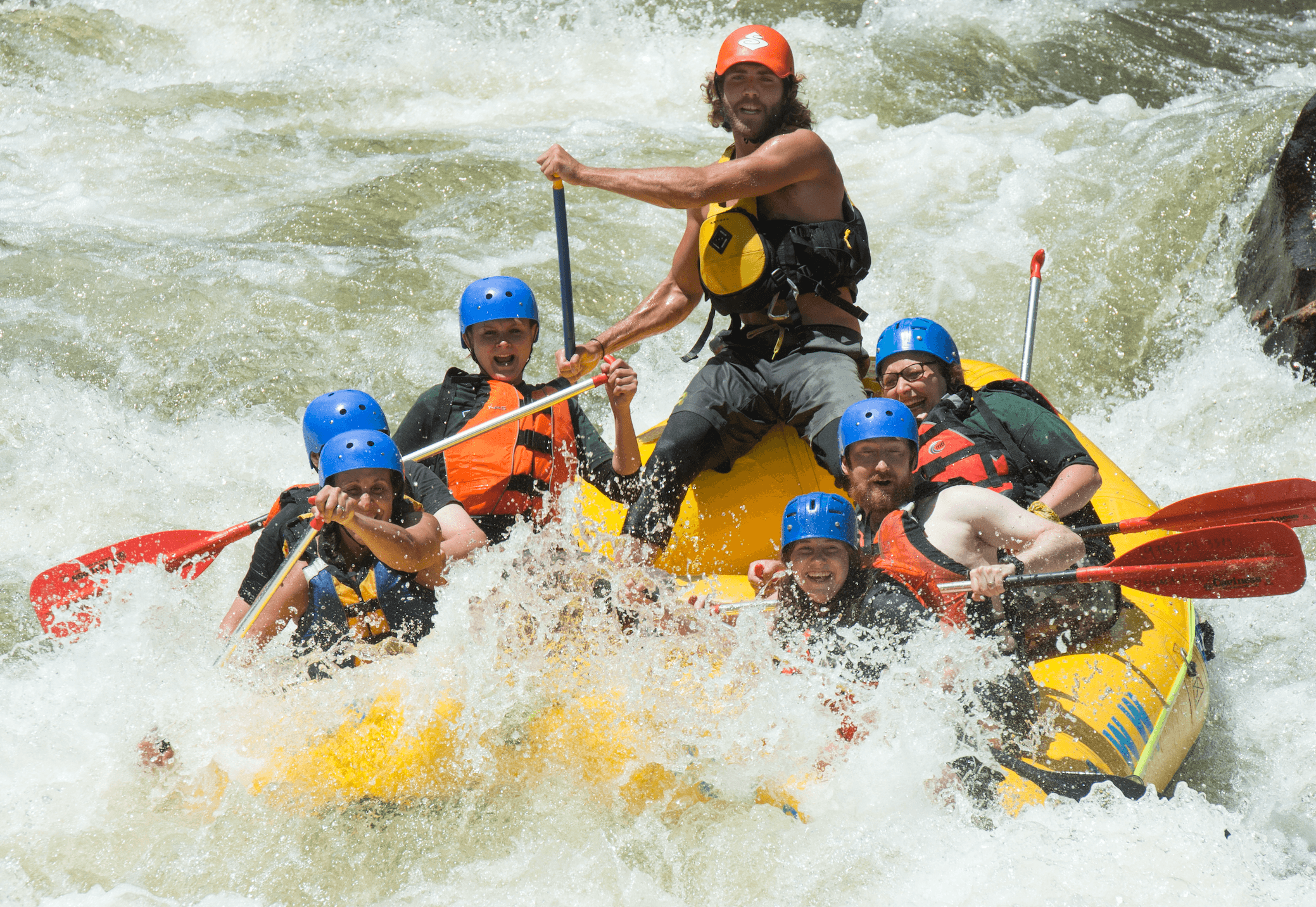 Six people are on a guided whitewater raft. They are wearing blue helmets and life jackets. Their guide stands in the back of the yellow inflatable raft wearing a red baseball cap. The water is white surrounding them.
