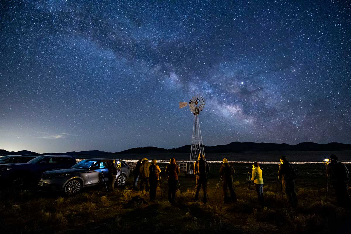 Two parked cars with six people standing next to them look up at the night's sky that's covered in tiny white stars. In the distance dark mountains look on at an old-school windmill.