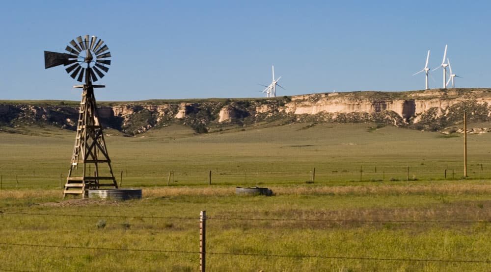 An old-school windmill turns in the foreground, and across some fences and prairie land, there are several white wind turbines