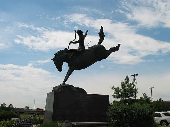 A life-size bronze sculpture of a bronco rider atop a horse stands in relief against a bright-blue sky peppered with white, fluffy clouds.
