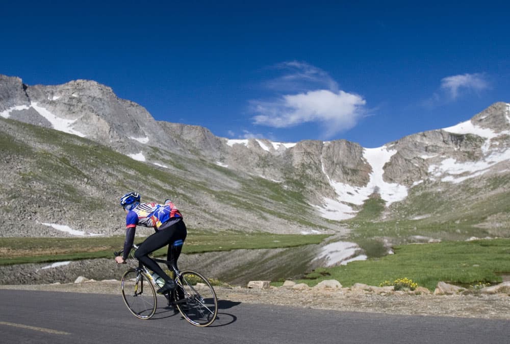 A cyclist zooms up a roadway under bright blue skies and a grassy meadow and a mountian ridge with a trace of snow